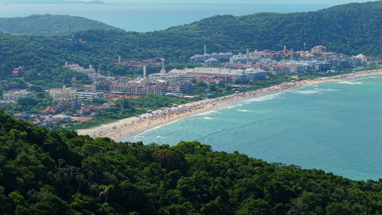 Pantai Praia Brava yang ramai di Florianópolis, dikelilingi oleh perbukitan berhutan dari sistem pegunungan pesisir Serra do Mar di Santa Catarina, Brasil, terlihat dari drone yang meluncur pelan.