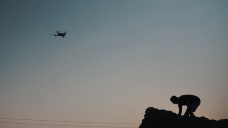 Silhouette-Des-Mannes-Auf-Einer-Klippe-Mit-Drohne,-Die-Während-Des-Sonnenuntergangs-In-Zeitlupe-Fliegt,-Krabi-Thailand