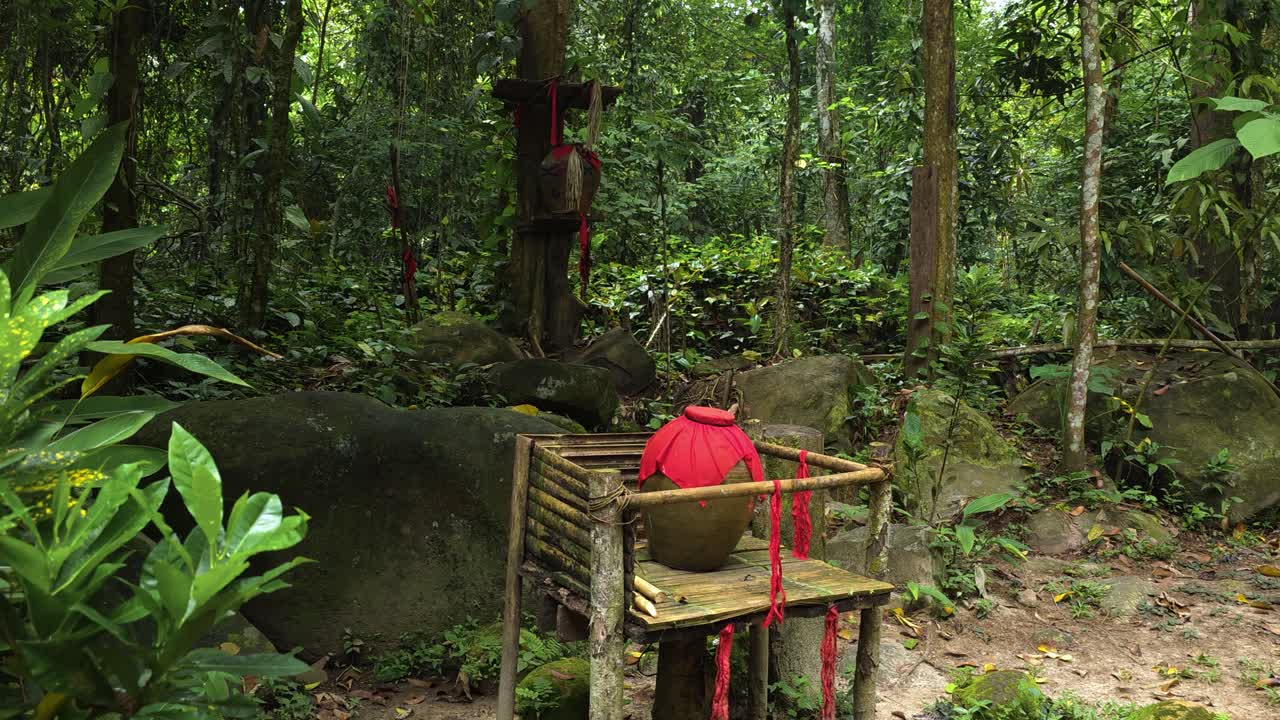 A traditional large red pot decorated with ribbons sits on a rustic wooden platform in a forest setting in Mari Mari cultural village, Kota Kinabalu, Malaysia