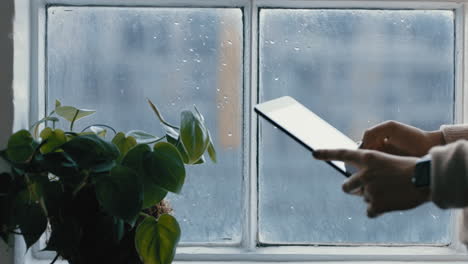 cerca de las manos de la mujer usando una tableta digital navegando por mensajes en línea leyendo redes sociales disfrutando de un dispositivo móvil con pantalla táctil de pie junto a la ventana relajándose en casa en un día frío y lluvioso
