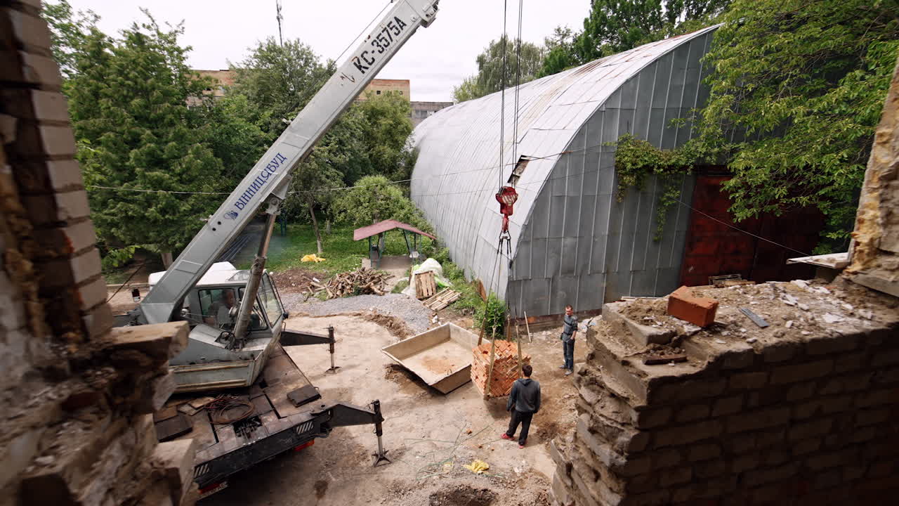 Looking from the higher floor of the constructed building into the yard. Men watch as the excavator lifting a pile of bricks.