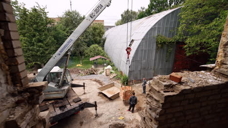 looking from the higher floor of the constructed building into the yard. men watch as the excavator lifting a pile of bricks.