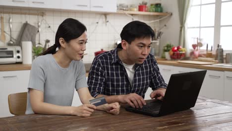 happy asian married couple enjoying shopping online with credit card and laptop. the man is keying in the number to make payment as the woman is reading to him