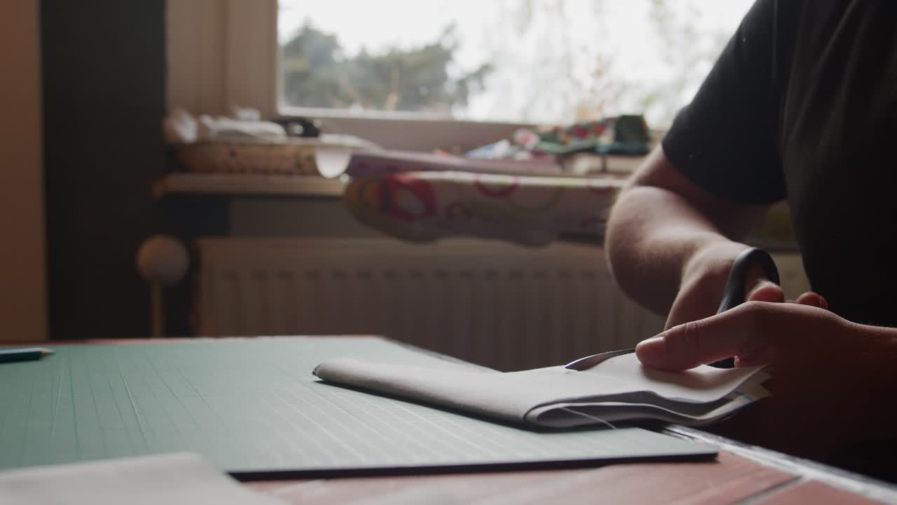 Seated woman cuts folded fabric in front of a window with craft materials in view