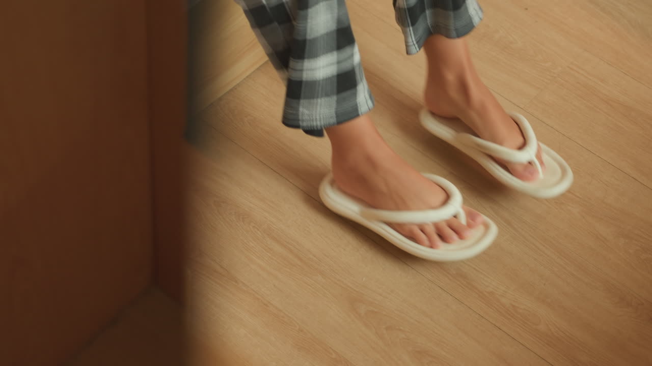 Rejsende tager hotelsko på ved døråbningen, hurtig fodbevægelse ind i plyssede hjemmesko på laminatgulv, aftenskygge og varmt indvendigt lys, ternet pyjamasærme og nærbillede antyder afrejsescene