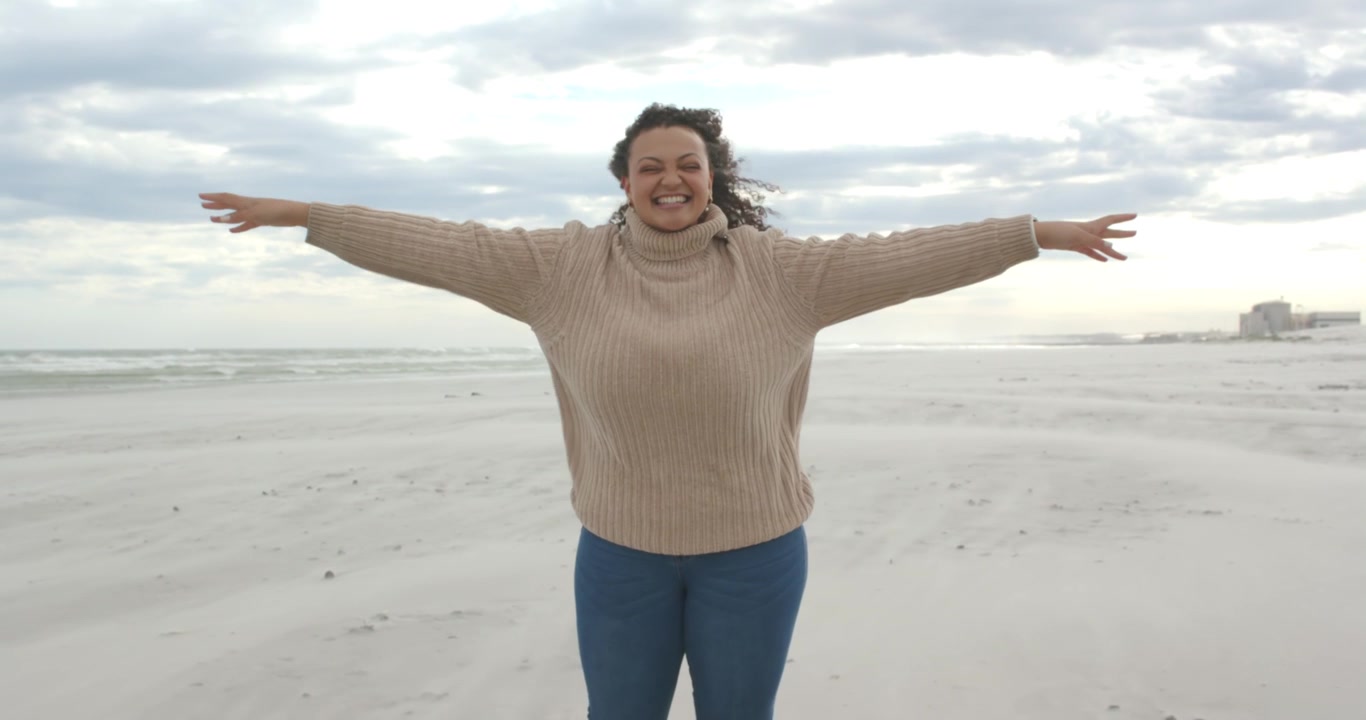 Mulher afro-americana com os braços abertos, o vento soprando, de blusa de gola alta bege e jeans na praia, com microfone.