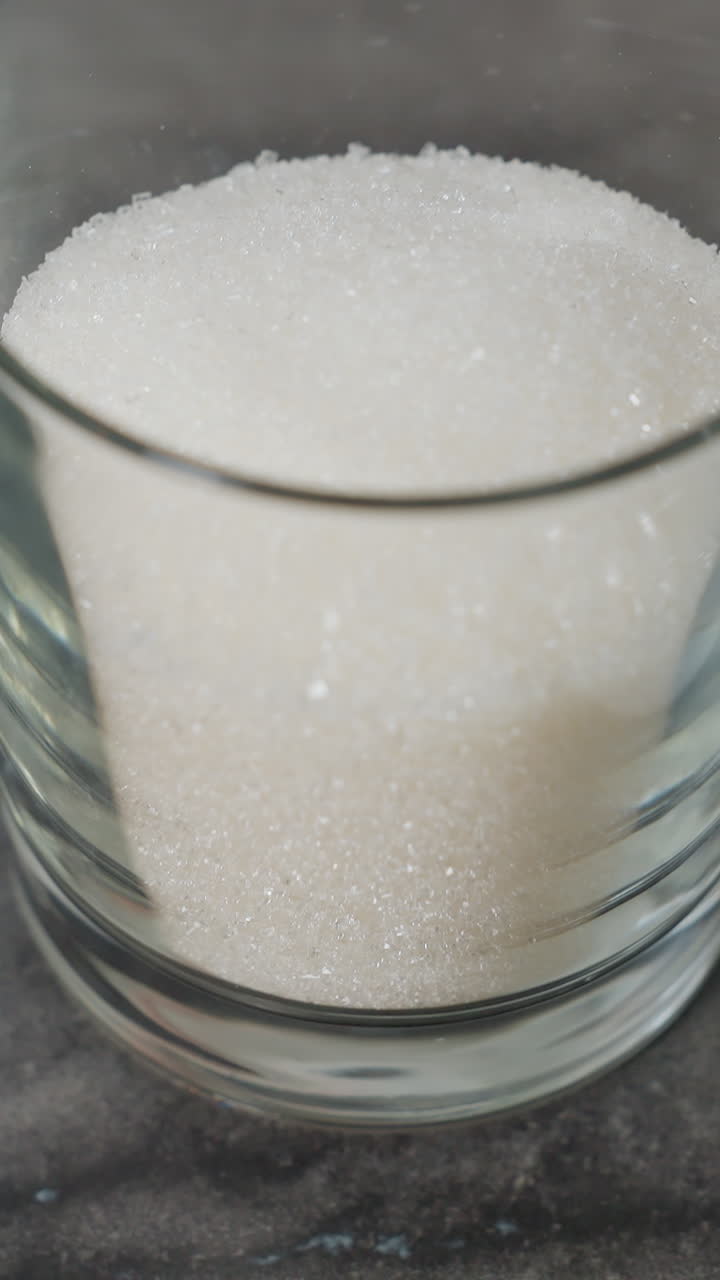 색 설탕으로 가득 찬 투명한 유리를 매끄러운 검은 테이블 위에 놓고 있는 손의 클로즈업, 나무 트레이와 주방 장비의 부분적인 시각