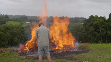 a man in a beanie stands in front of a raging bonfire on a bush property