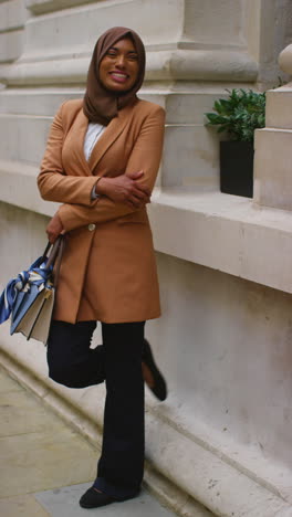 vertical video portrait of smiling muslim businesswoman wearing hijab and modern business suit standing outside city office buildings 8