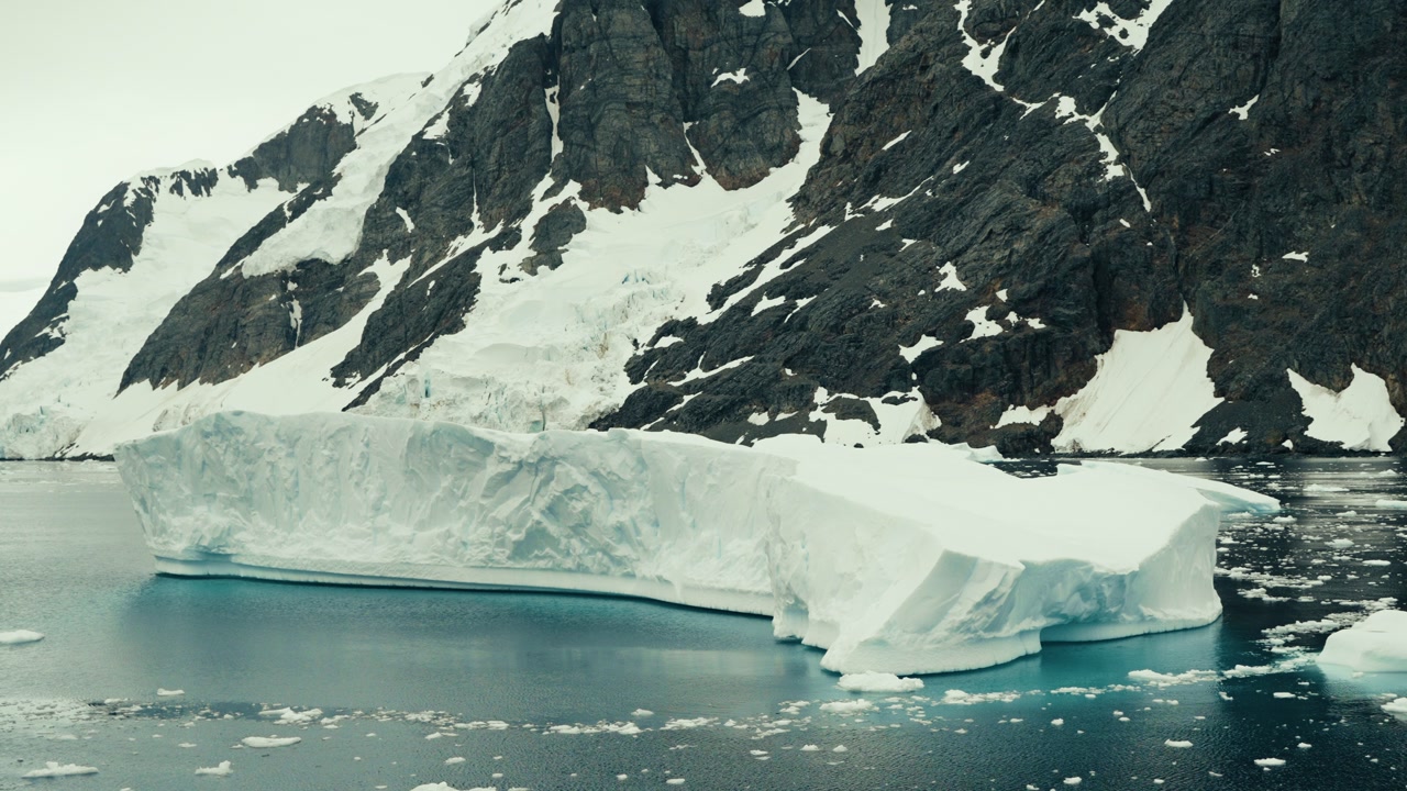Massive tabular iceberg drifts through glassy teal waters of Lemaire Channel with dramatic snow-covered rocky cliffs rising behind in slow cinematic parallax shot