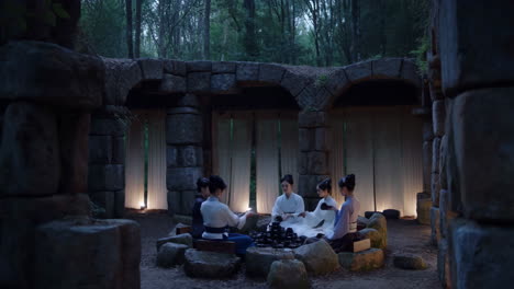 Women in traditional attire playing a board game in a serene forest setting