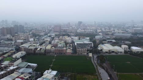 A-dense-urban-area-with-buildings-and-a-green-field-on-an-overcast-day,-misty-atmosphere,-aerial-view