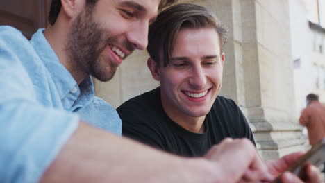 Una-Pareja-Gay-Masculina-Sentada-Al-Aire-Libre-En-Las-Escaleras-Del-Edificio-Mirando-Teléfonos-Móviles