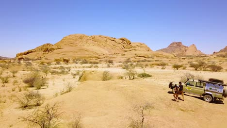 Aerial-over-a-couple-walking-near-a-4WD-safari-jeep-in-the-rugged-desert-landscape-of-Spitzkoppe-Namibia-Africa