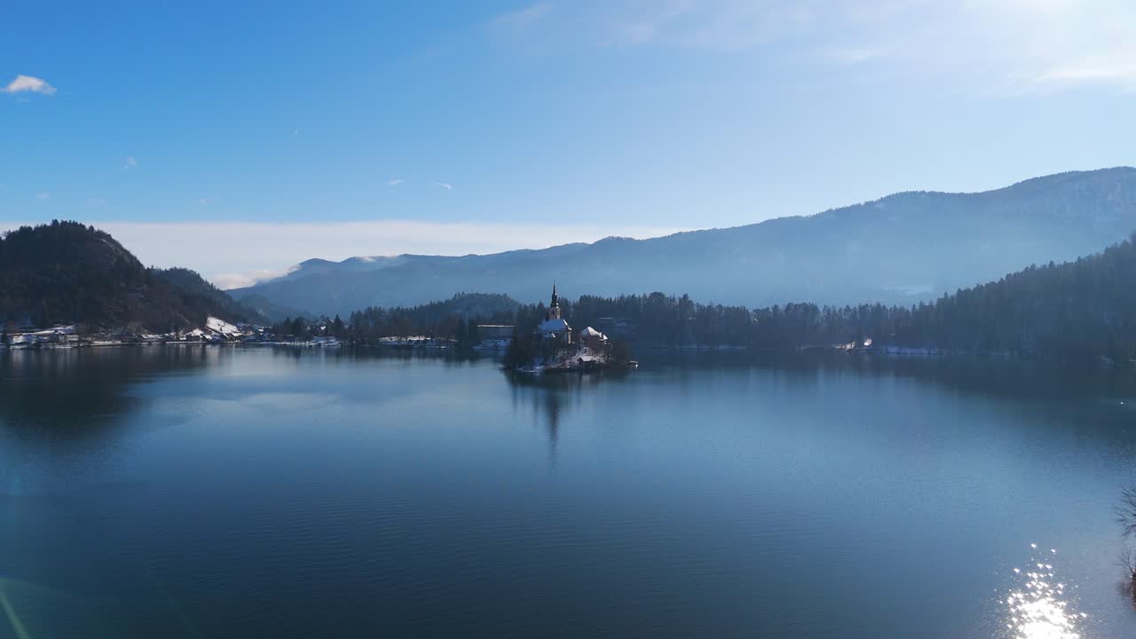 Ampia vista invernale del Lago di Bled con l'Isola di Bled e la sua chiesa al centro di acque calme e blu, incorniciato da colline innevate e distanti montagne slovene