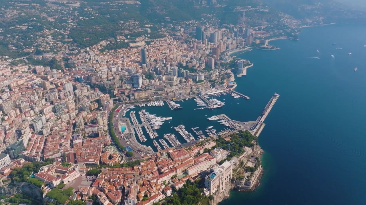 La costa e il porto di Monaco. La telecamera riprende yacht ed edifici lungo il lungomare. L'ambientazione evidenzia la vivace attività della zona.