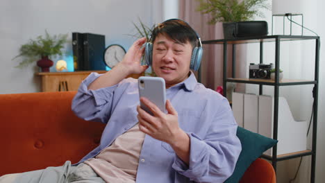 happy asian man in wireless headphones relaxing sitting on sofa at home listening favorite music