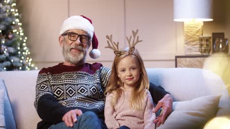 portrait of loving happy senior grandpa in glasses wearing santa hat spending christmas together with joyful grandchild sitting in decorated room near glowing xmas tree embracing together