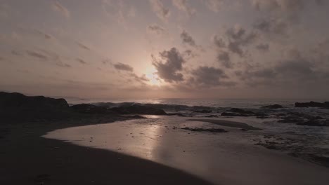 Aerial-view-low-flying-rocky-sandy-sunset-beach-with-dramatic-cloudscape-horizon