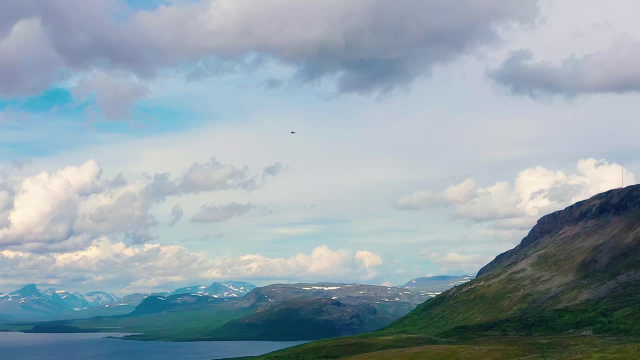 Aerial view tracking a helicopter flying over the Saanatunturi mountain, lake Kilpisjarvi and polar nature, cloudy, summer day, in Lapland, Finland - pan, drone shot