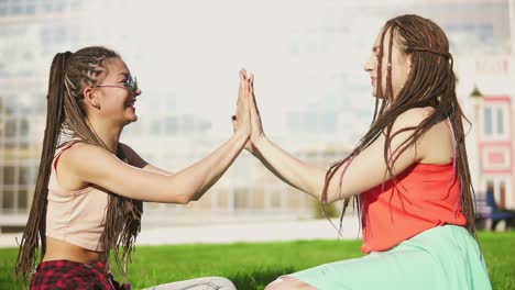 happy women with dreads sitting on grass in summer park. young friends talking and playing outdoors having fun. mulatto woman