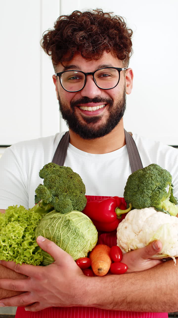 Uomo con grembiule rosso che tiene verdure fresche in primo piano, per la preparazione di pasti sani e nutrienti nella cucina di casa.