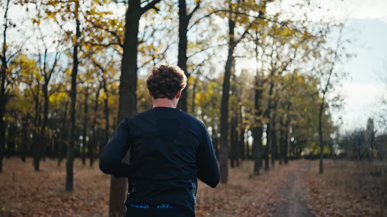 vista trasera de un hombre moreno con un uniforme deportivo negro con cabello rizado y una barba corre a lo largo de un camino de tierra durante una carrera matutina en el bosque soleado de otoño