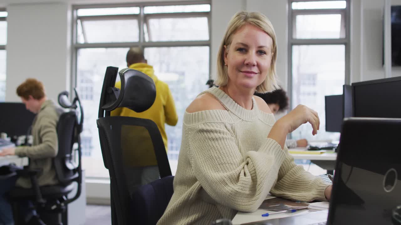 Porträt einer lächelnden, kaukasischen Kreativunternehmerin, die an ihrem Schreibtisch in einem modernen Büro sitzt.