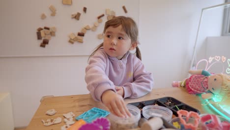young toddler girl with curious interested expression explores options of toys to play with in an indoor playground art station, full of wonder, frontal medium static, slow motion