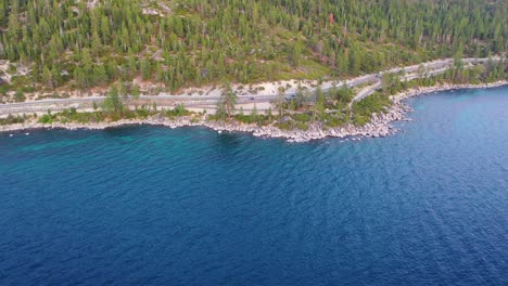 Aerial-Drove-View-of-Lakeside-Road-with-Cars-Driving-at-Late-Taho-with-Pine-Tree-Forest,-Rocky-Riprap-Bank-and-Clear-Blue-Water-Along-Scenic-Coast-in-California,-USA