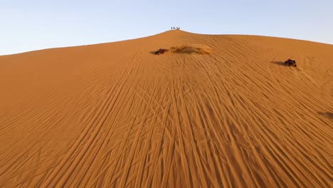 Buggies-racing-toward-people-up-a-golden-sand-dune,-aerial-push-in