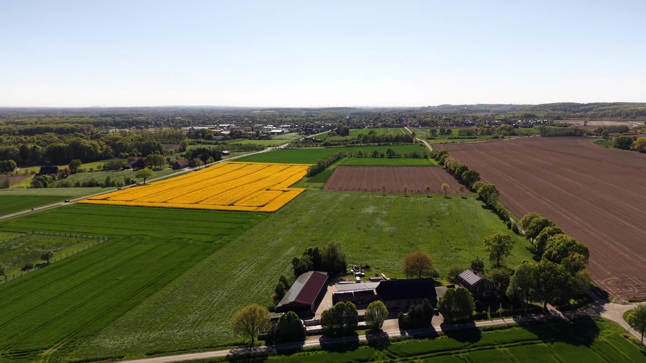 Farbenfrohe Agrarlandschaft in Europa mit Rapsfeldern, grünen Wiesen und einem ländlichen Gehöft unter hellem Frühlingssonnenlicht, die landwirtschaftliche Muster und eine friedliche ländliche Szenerie zeigt. Luftaufnahme.