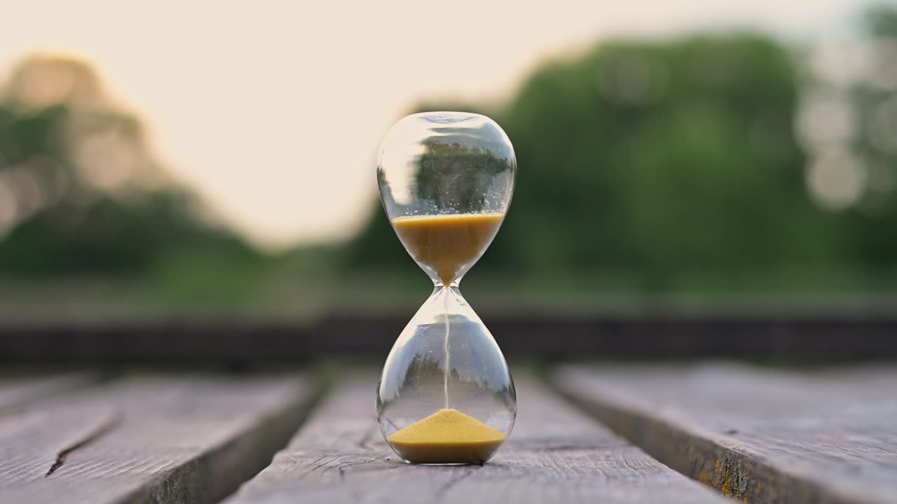 A glass hourglass filled with golden sand sits centered on a rustic wooden platform outdoors. The sand flows steadily as soft evening light and a blurred green background create a serene atmosphere.