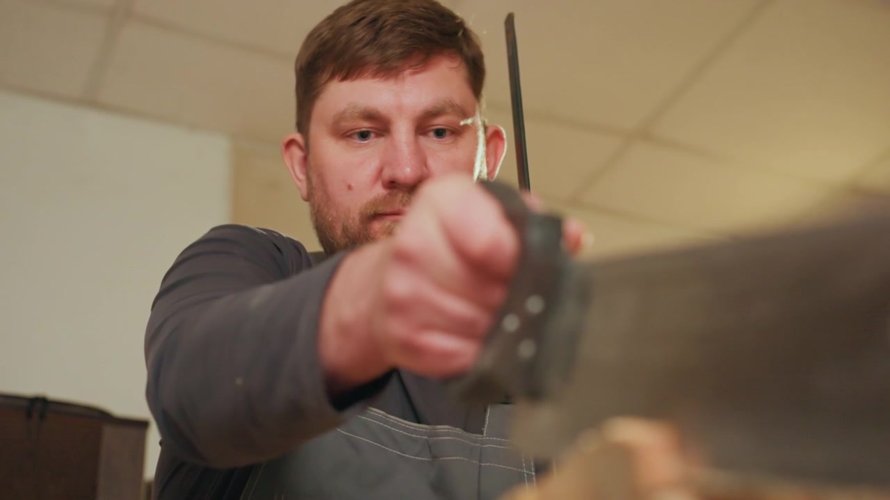 Close up shows focused expression and steady hands of craftsman as man operates large metal hand saw cutting through thick wood in indoor workshop setting. background reveals plain walls