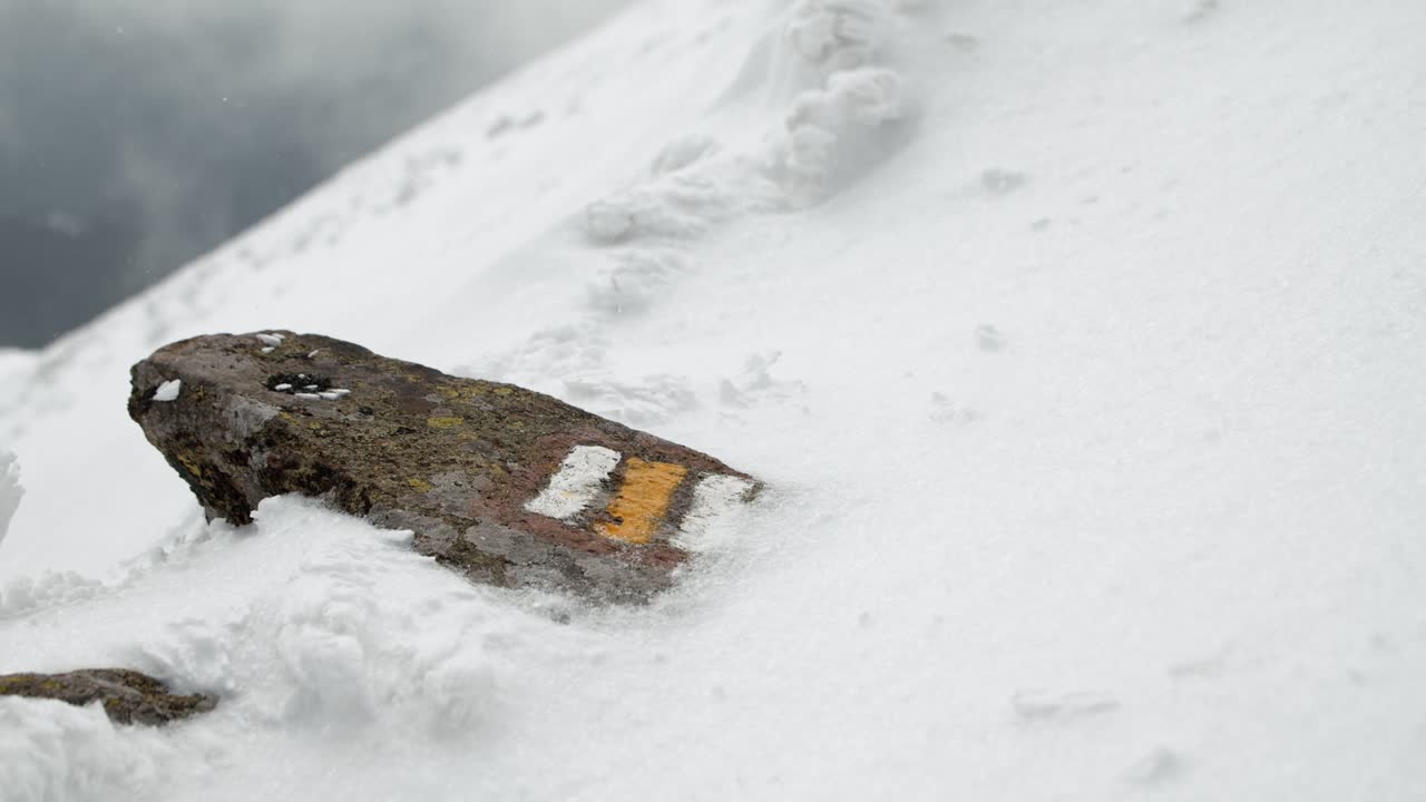 Close up of a yellow touristic route sign on a rock covered with snow on icy and snowy pathway during winter and snowfall