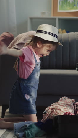 pretty preschooler girl with hat puts clothes pile into suitcase and imitates airplane at home. happy child prepares for first flight abroad