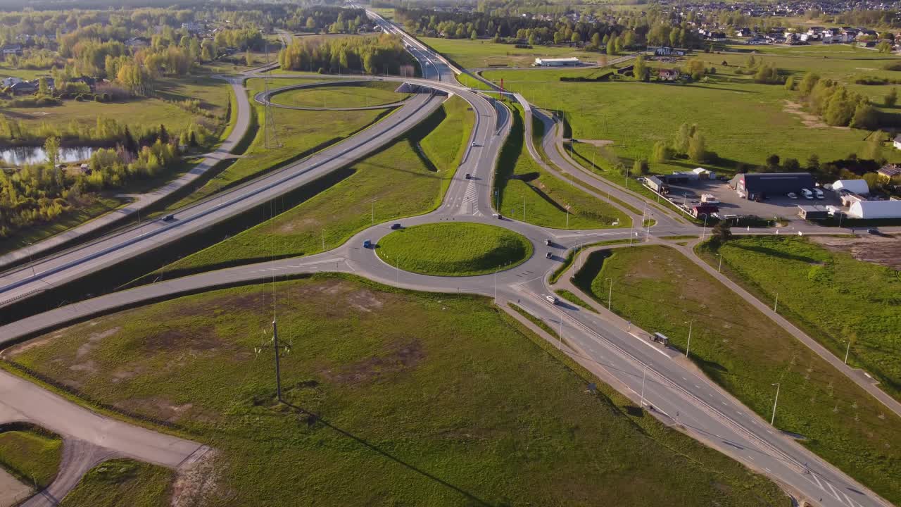 Aerial view of cars driving on roundabout and highway in Katlakalns residential area, Latvia during spring, surrounded by green fields and trees