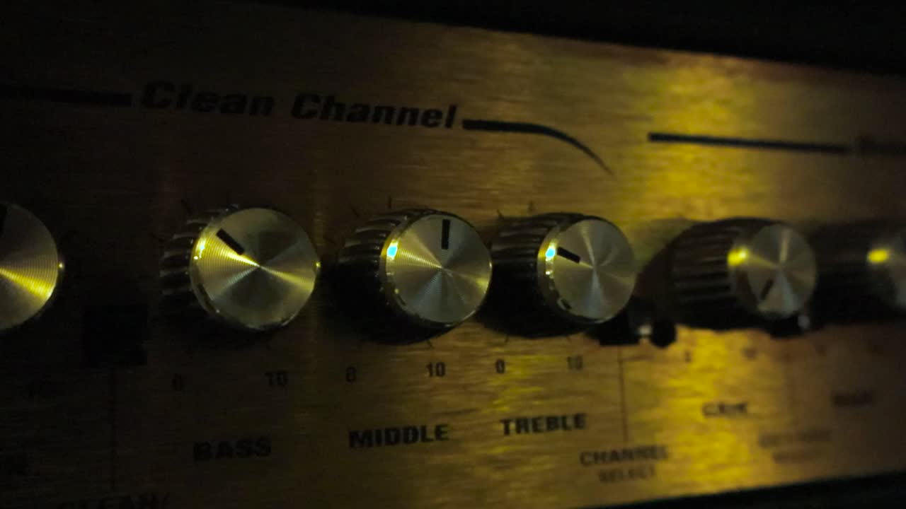 Moving sideways over electric guitar amplifier. Detail view of volume knobs in dark studio room and light glows on button panel creating warm metallic shine. Close up panning electronic device system