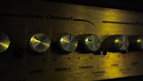 moving sideways over electric guitar amplifier. detail view of volume knobs in dark studio room and light glows on button panel creating warm metallic shine. close up panning electronic device system