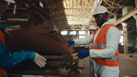 A-man-with-black-skin-and-a-beard-in-a-white-uniform-and-an-orange-vest-together-with-his-employees-sorts-waste-paper-at-a-waste-and-garbage-processing-plant