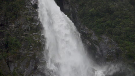 Cámara-Lenta-Bajando-Por-Una-Gran-Cascada-En-Un-Acantilado-Con-Un-Bosque-En-Primer-Plano---Milford-Sound,-Nueva-Zelanda