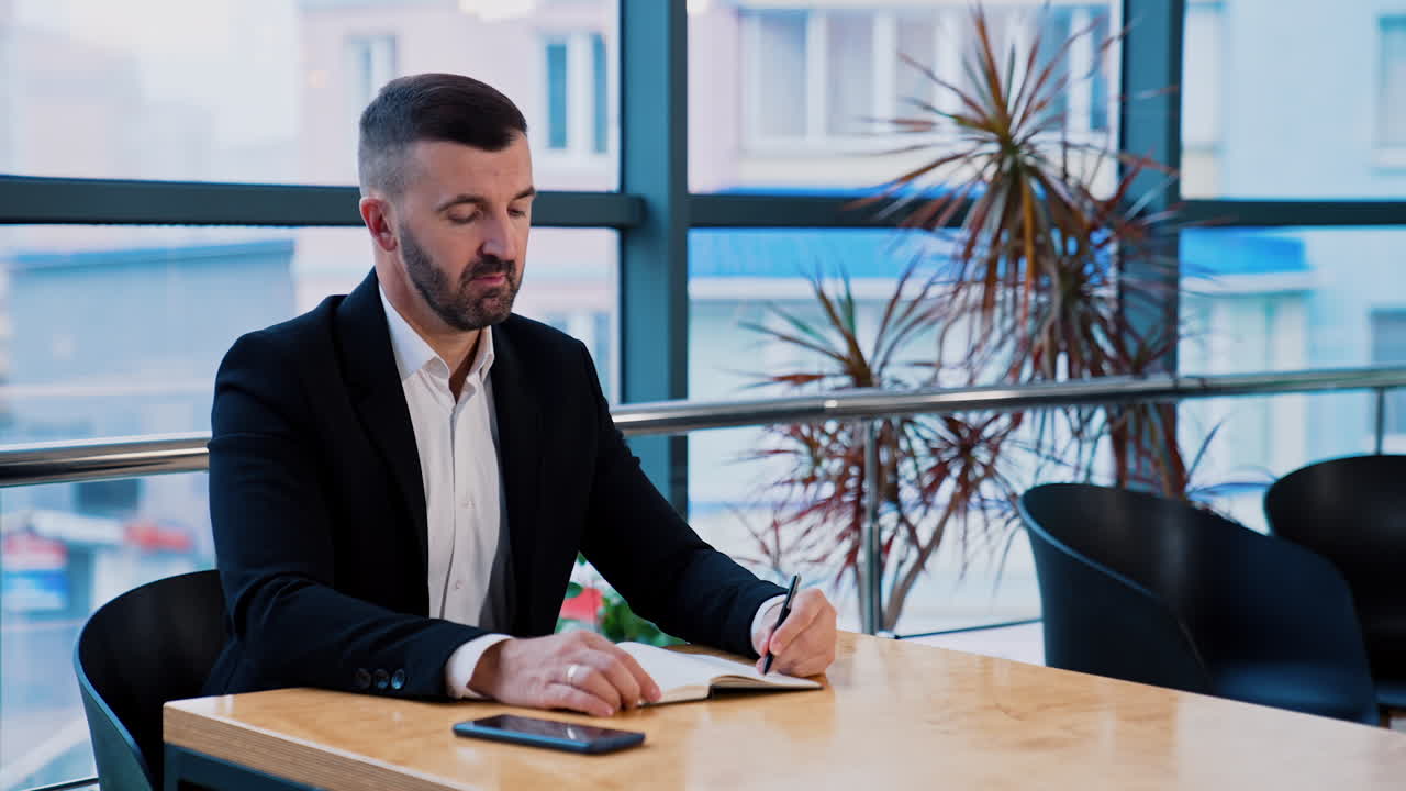 Busy boss writing something in paper notebook. Businessman takes his phone and scrolls information. Big windows at backdrop.