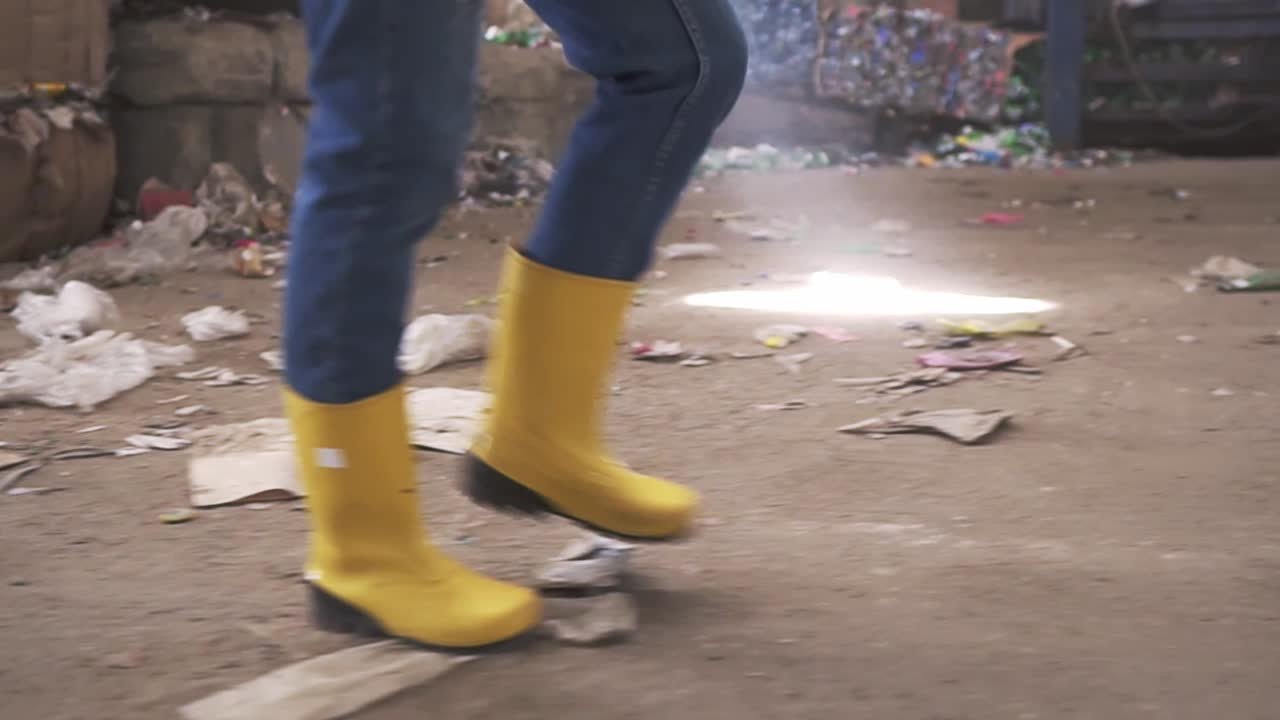 Unrecognizable woman in protective working clothes - yellow boots walking by waste recycling factory, huge area with used bottles and garbage. Close up footage