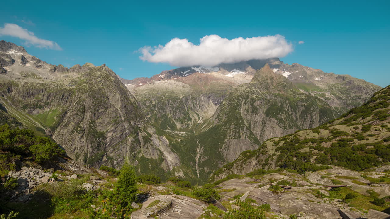 Majestic alpine mountains with rugged cliffs and green valleys beneath a clear blue sky. A cloud hovers over snow-capped peaks
