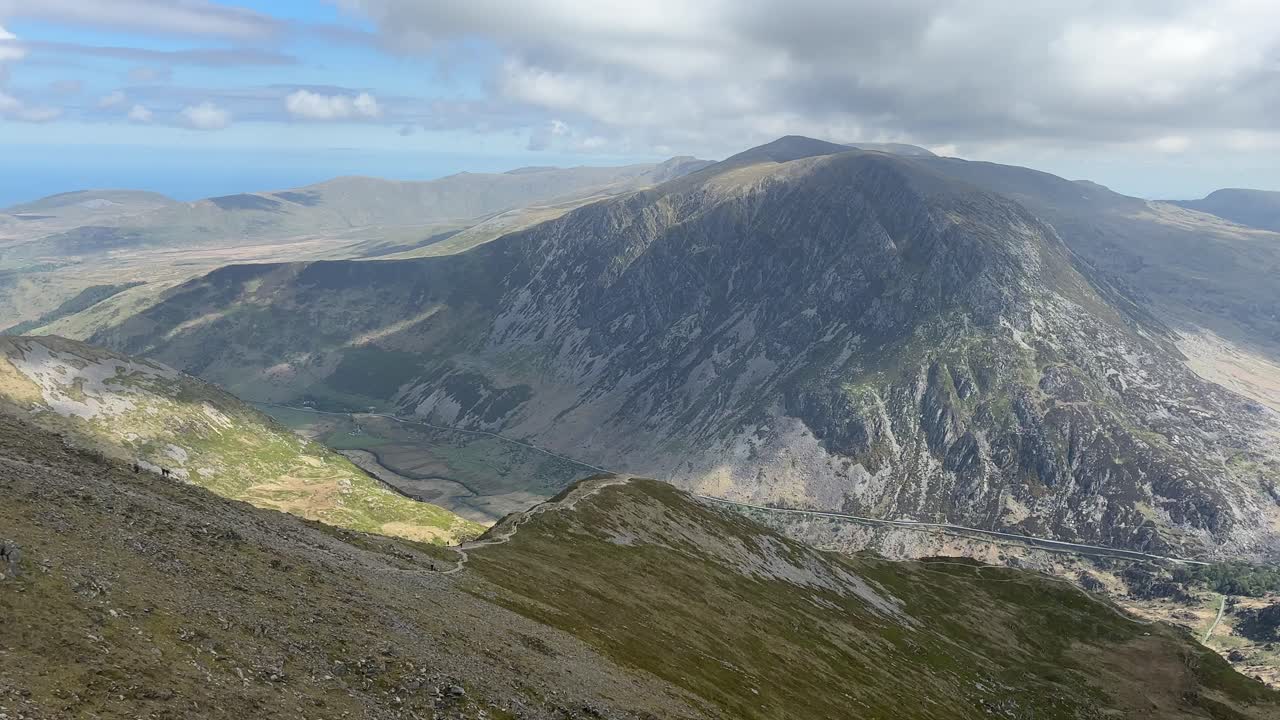 Expansive view from Y Garn summit in Snowdonia shows rugged cliffs, green valleys, and distant peaks under broken clouds, highlighting natural beauty and dramatic landscape of North Wales