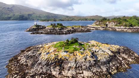 aerial drone view of rocky mossy island with white lighthouse amid blue water and highlands