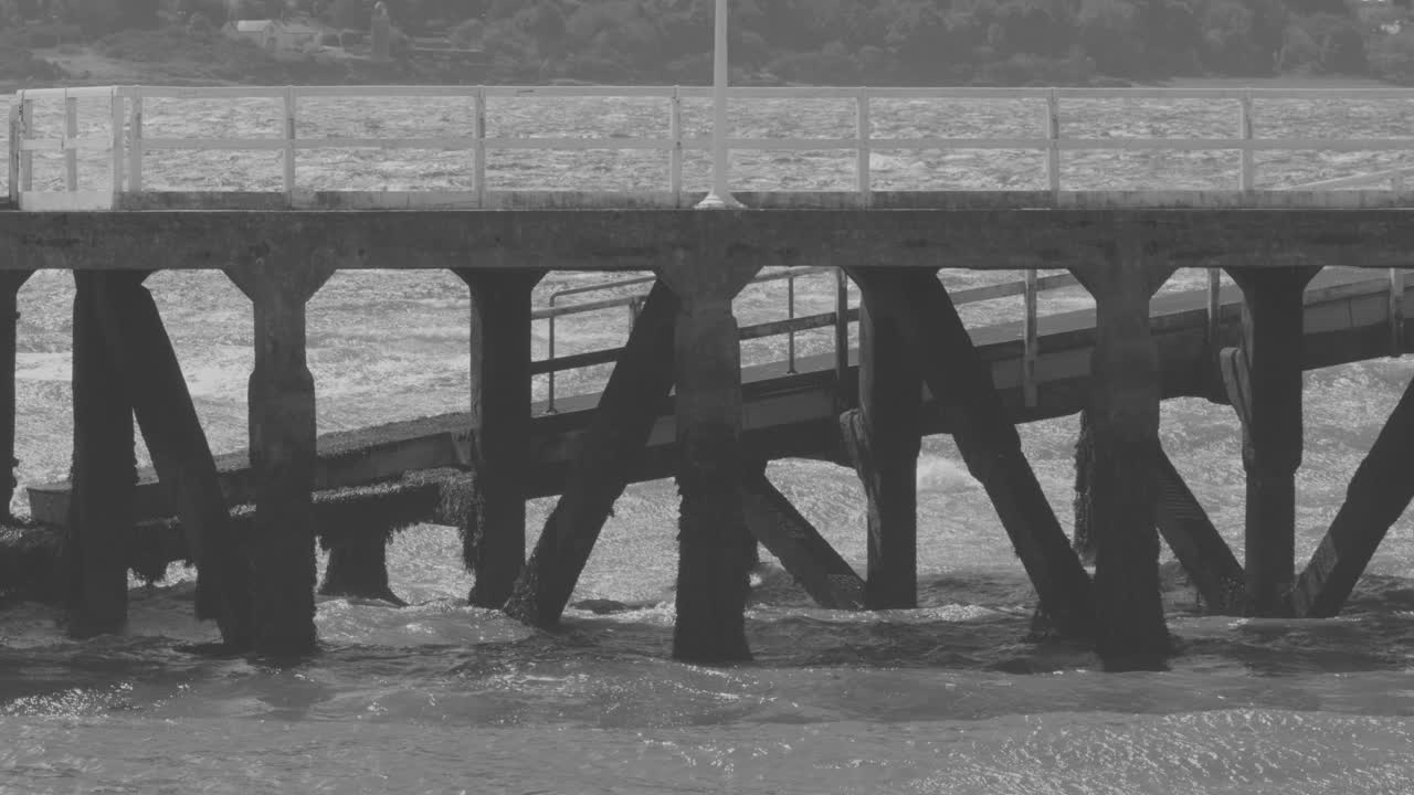 Grayscale video of wooden pier with waves, overcast lighting, steady camera, coastal Highlands environment