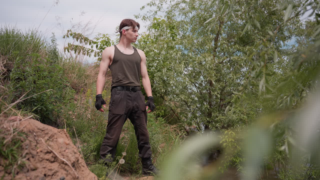 Adventurer wearing tank top, gloves, and cargo pants stands alert on slope by riverbank, scanning surroundings with focused gaze under cloudy daylight in lush green outdoor terrain with trees and soil