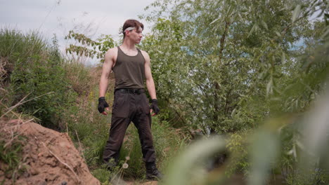 adventurer wearing tank top, gloves, and cargo pants stands alert on slope by riverbank, scanning surroundings with focused gaze under cloudy daylight in lush green outdoor terrain with trees and soil