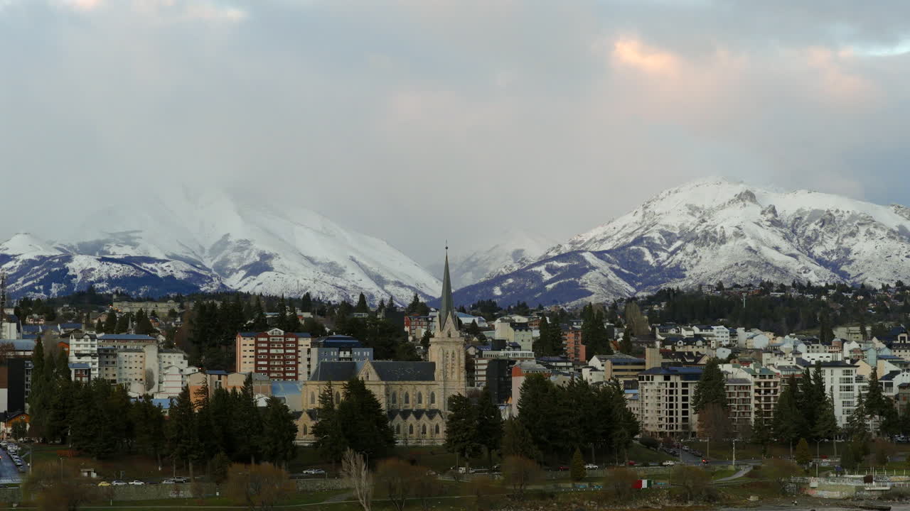 Drone descends at Bariloche, framing historic church and buildings with snow-capped mountains behind